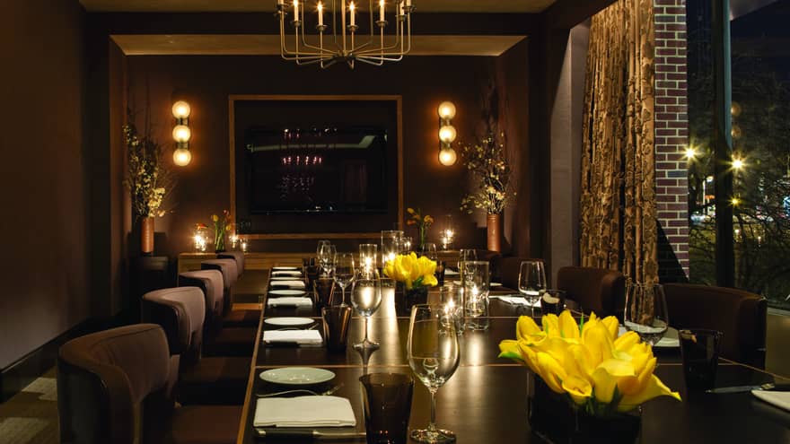Long private dining table with plush chairs set with wine glasses, yellow flowers, candles, low lights on walls