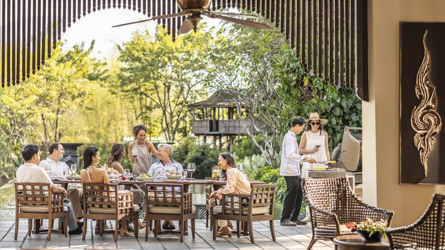 Group sharing a meal and drinks on an outdoor terrace with a chef grilling nearby, surrounded by lush greenery.