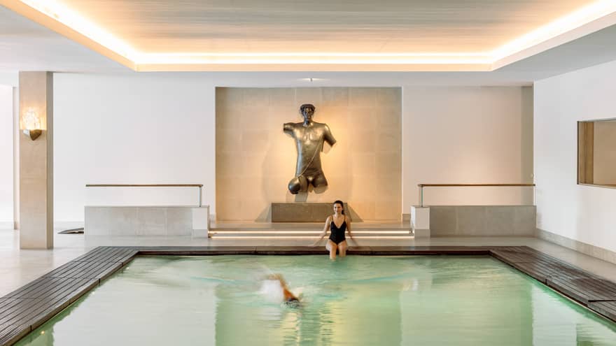 Indoor pool with a swimmer and a person sitting at the edge, featuring a large bronze torso sculpture on the far wall.