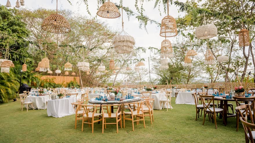 A lawn is set for an outdoor party with various round tables, white table cloths and a variety of straw light fixtures hanging from vined ropes overhead