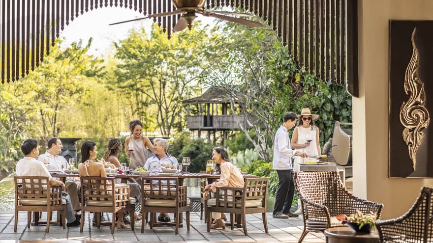 Group sharing a meal and drinks on an outdoor terrace with a chef grilling nearby, surrounded by lush greenery.