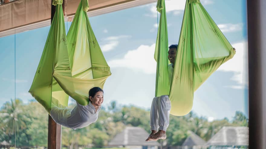 Two people practising antigravity yoga in bright green hammocks, suspended in a wellness resort studio with a view of lush tropical surroundings