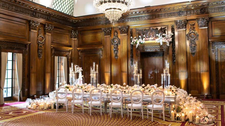 Elegant wedding reception setup in a grand ballroom adorned with chandeliers and a floral-covered table, showcasing regal decor