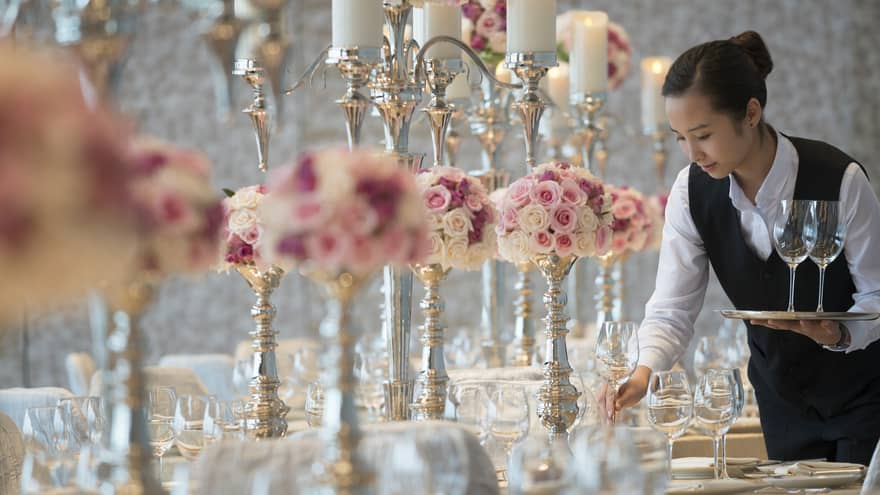 Hotel staff sets wine glasses on Peony Ballroom banquet table with pink flowers, tall candles