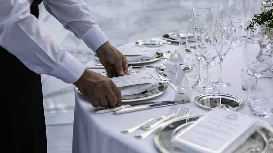 A server in a white shirt sets a formal dining table, placing a menu on a silver plate. The table is elegantly arranged with glassware, silverware and name cards.