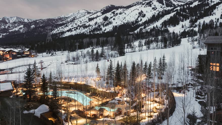 Aerial view of illuminated blue outdoor swimming pool through trees on snowy hill at dusk