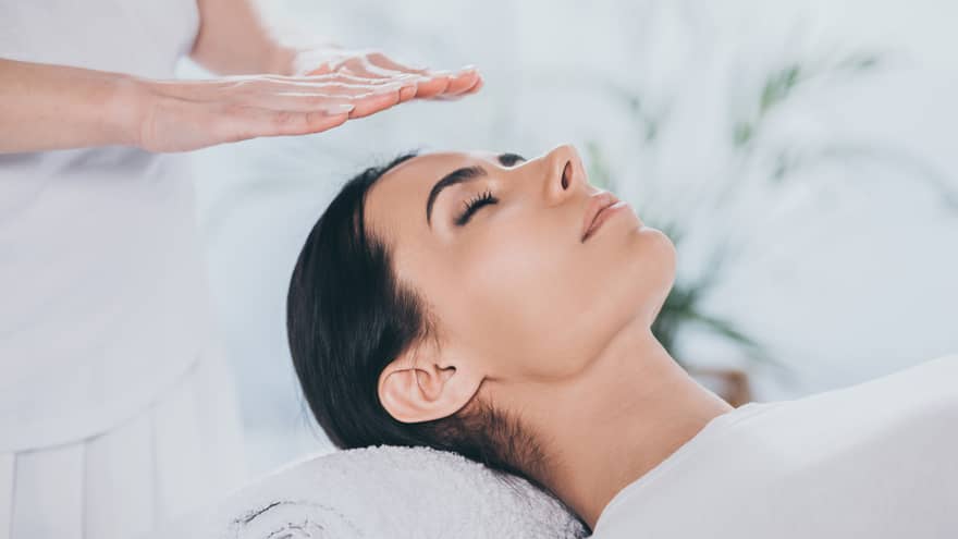 Woman with eyes closed lays on rolled up towel on spa table under therapists' hovering hands