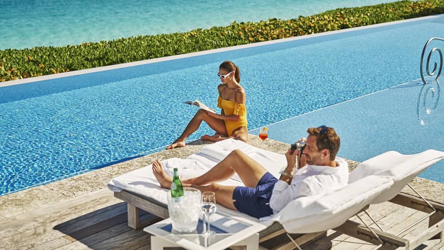 Two people relaxing by a pool, one reading a book in an orange swimsuit and sunglasses, the other lounging on a chair with a camera