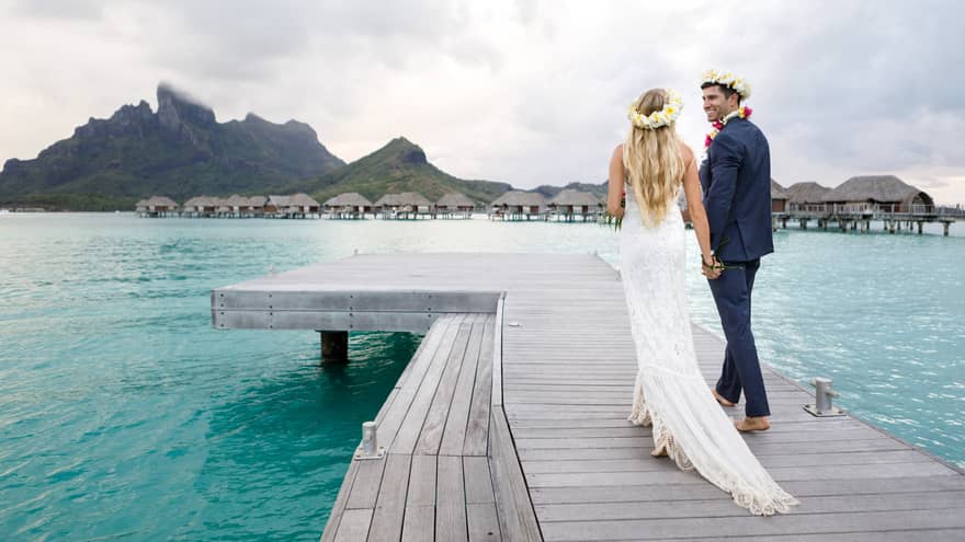 Back of groom and bride with long trail walking on dock over lagoon