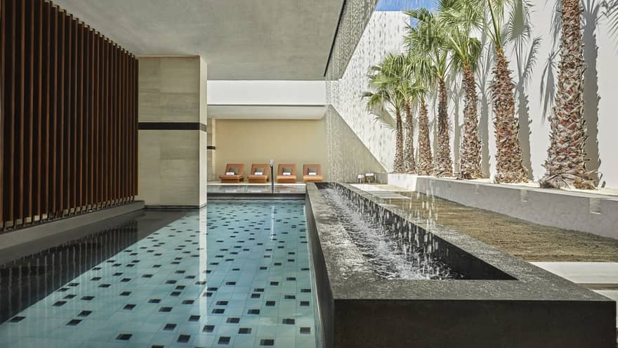 An outdoor pool is covered with a white stucco overhang and lined with lounge chairs on one side, palm trees on the other