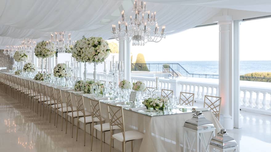 Long elegant wedding banquet table on patio with chandeliers, white floral arrangements