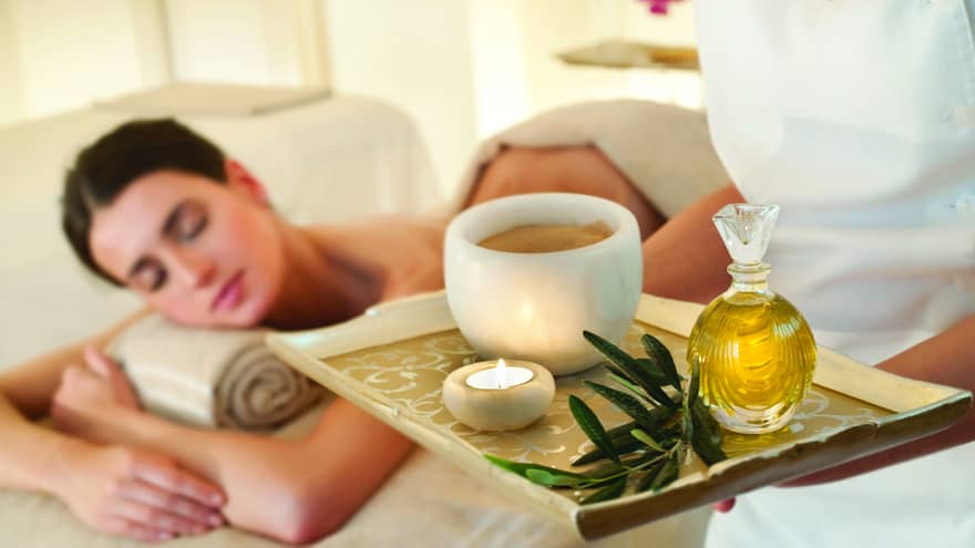 Spa staff holds tray with tealight candle, perfume, marble bowl near woman lying on massage table