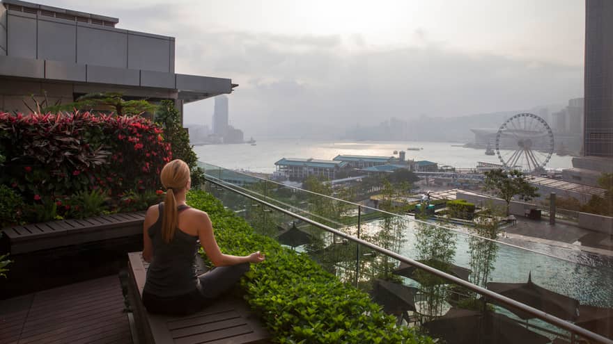 Back view of woman meditating on wood bench on deck high above hotel pool, patio, harbour