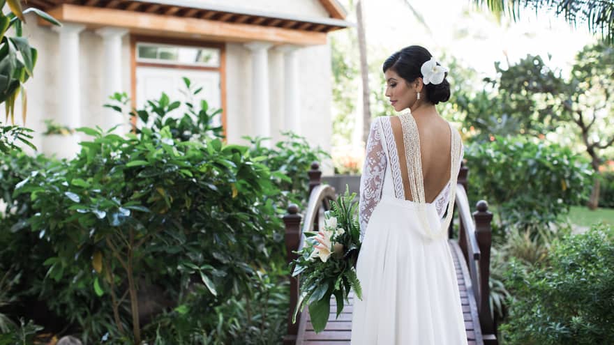 A stunning bride wearing a white wedding gown and flower in hair, gracefully poses on a charming wooden bridge in a lush garden