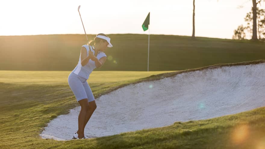 Woman swings golf club in sand trap on green at sunrise