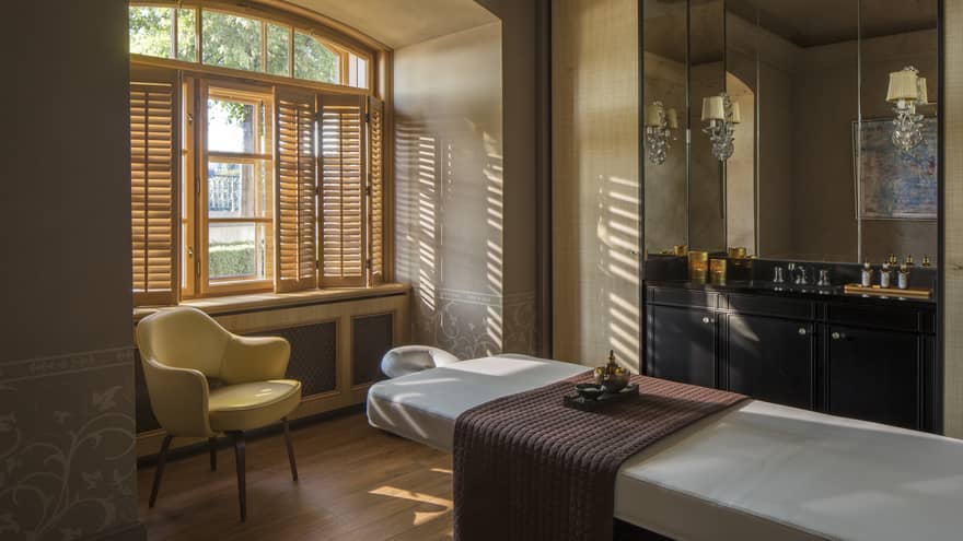 White massage table with quilted brown blanket, mid-century style armchair in front of sunny window, open shutters