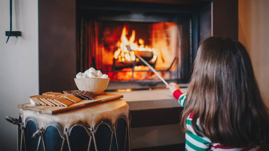 A little girl in red and green pajamas roasts marshmallows in a fireplace