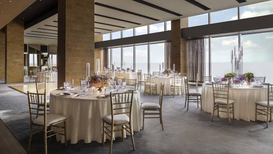 A wedding banquet hall featuring round tables and chairs, set with cream and gold decor, long glass candle holders, and floral centrepieces.