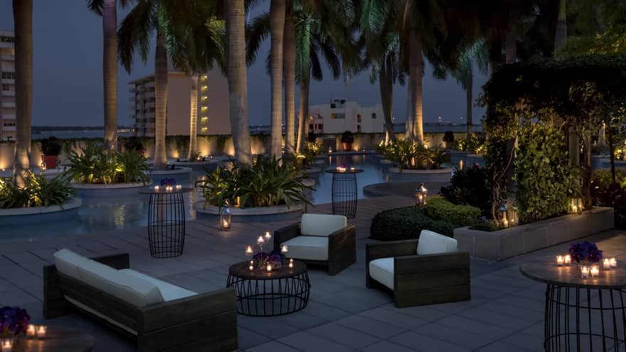 Plush white patio sofas and chairs, candle-lit tables under palm trees at night