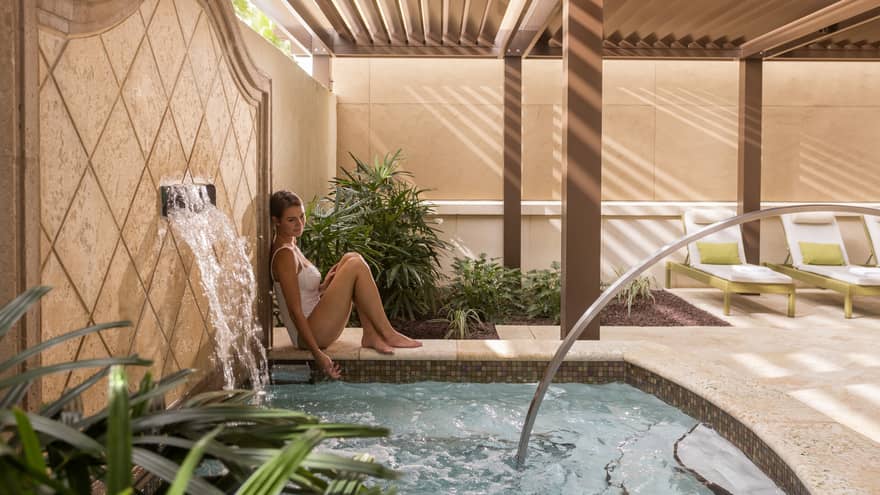 Woman wearing white bathing suit sits next to a whirlpool with a water fountain