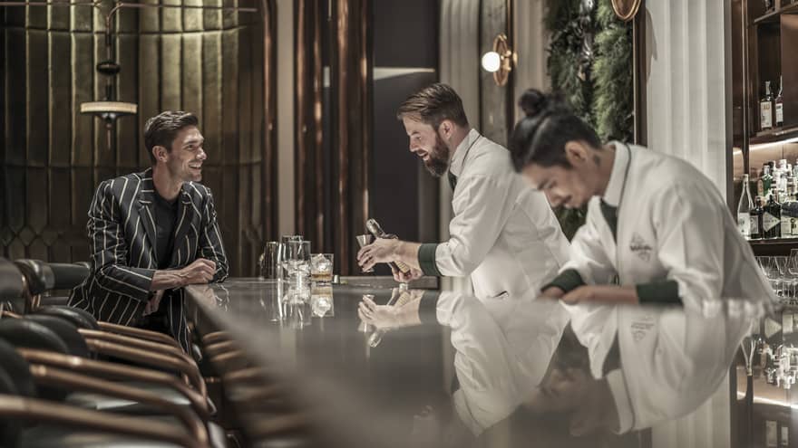Bartender preparing a drink while a customer sits at the bar, both smiling, in a modern, upscale bar with a reflective countertop and warm lighting.