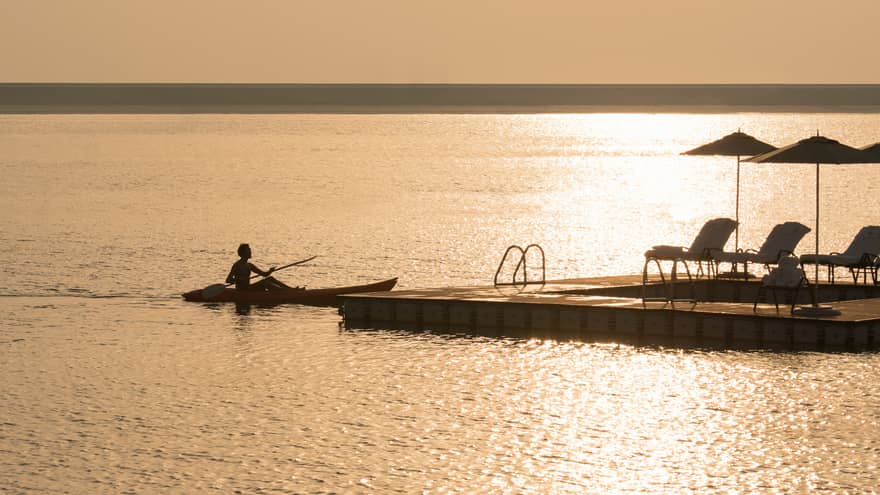 Silhouette of person in kayak with paddle on water in front of dock with patio chairs, umbrellas at sunset