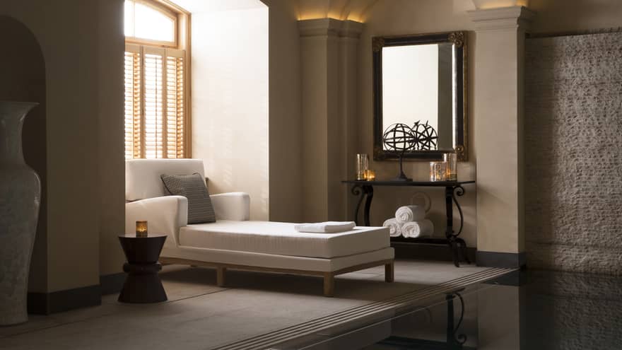 Long white chaise with accent pillow under sunny spa window with shutters, candles on shelves in room