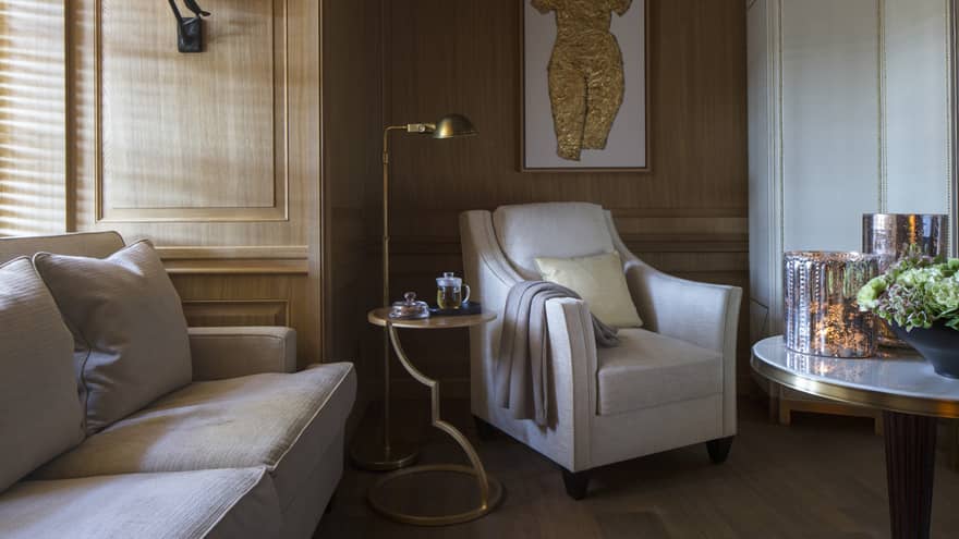 Sunlight streams through window to corner of sofa, white armchair with blanket on arm, reading lamp, art piece on wall