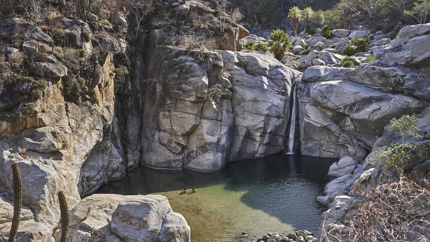 Two people wade in a small ocean lagoon surrounded by large cliffs, creating a serene and picturesque scene with clear water and rugged natural beauty.