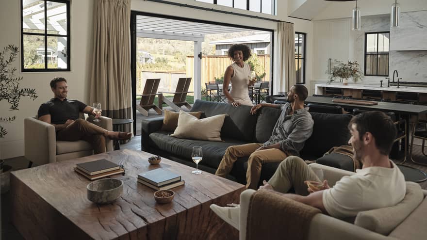 Friends gather around a table in a living room, seated on a couch and club chairs, with a view of the villa's outdoor area.