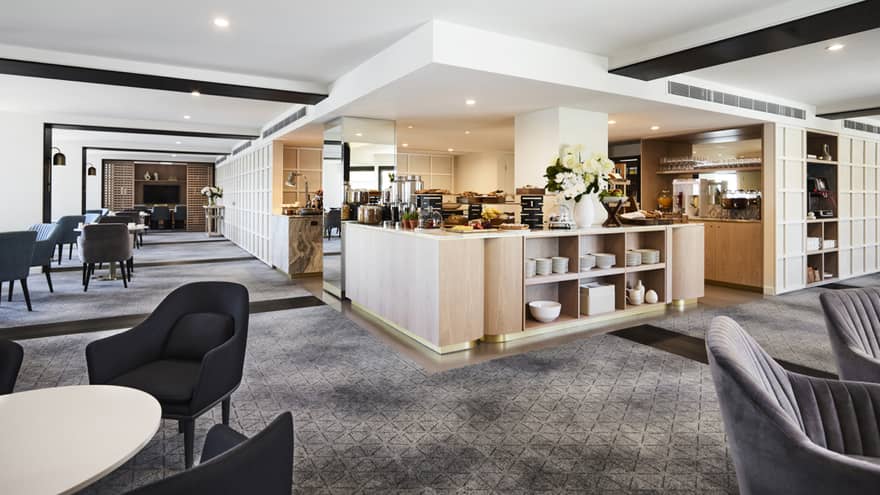 A modern hotel lounge with plush grey chairs and carpet, and a well-equipped kitchen set up with buffet foods and coffees.