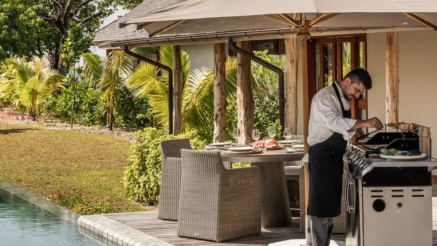 Chef wearing apron cooks at barbecue on deck by dining table, pool