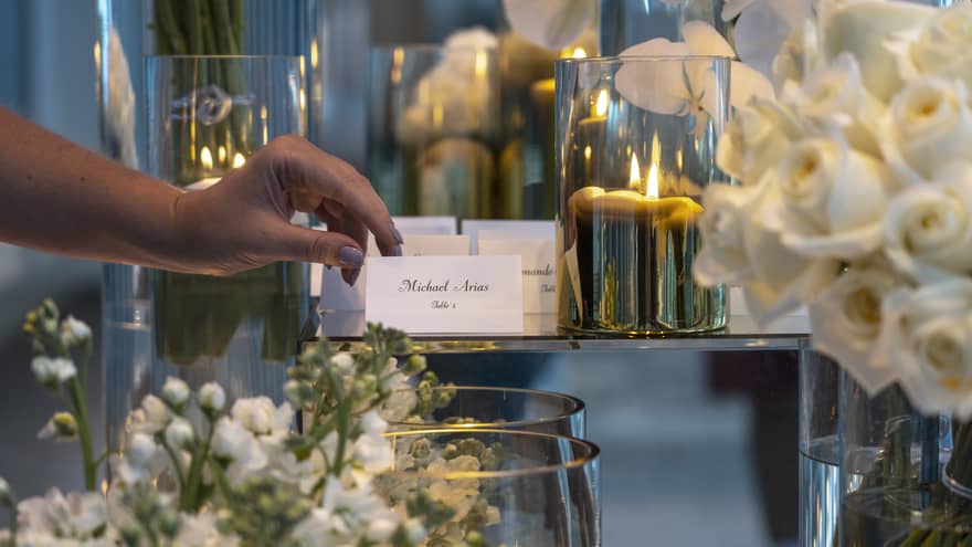 A close-up of a wedding guest picking up a place card from an elegant display at Four Seasons. The table features a sophisticated arrangement of white roses, with golden candle votives softly illuminating the background, creating a warm and welcoming ambiance for the celebration.