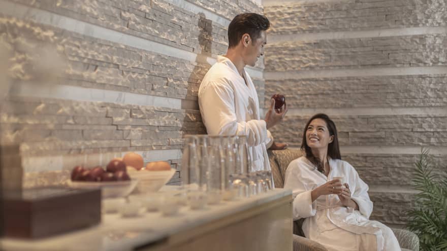 Two people in white robes relax in a spa lounge, one holding an apple. Various fruits and glasses sit on a wooden surface. The atmosphere is calm and inviting.