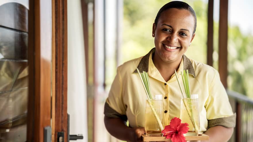 Smiling residence villa host in uniform holds tray with two cocktails, red tropical flower
