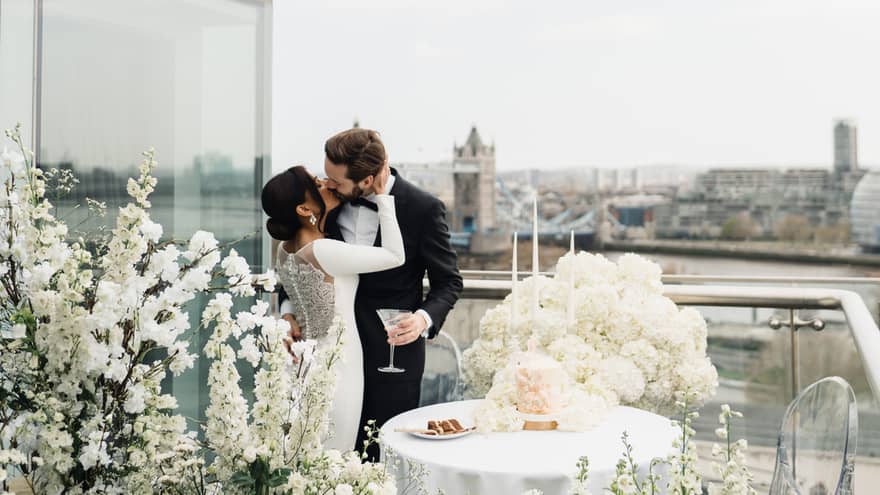 A bride and groom share a kiss on a flower-adorned balcony with stunning views of the city in the background.