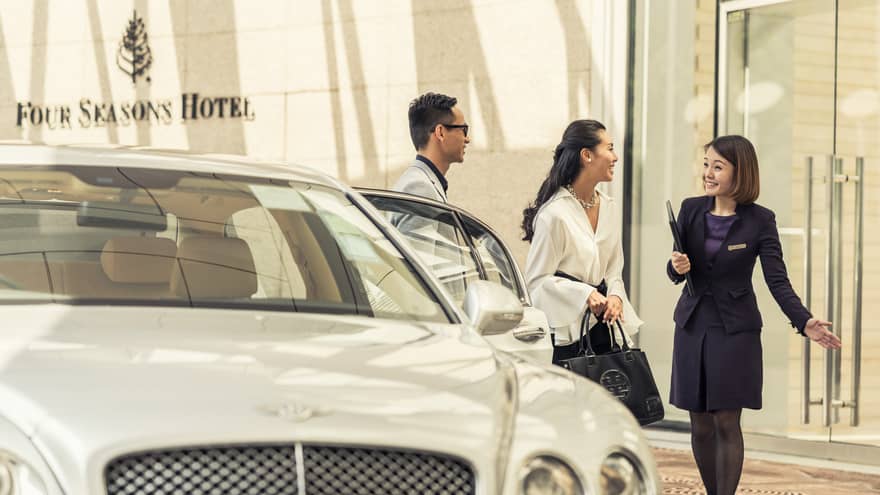 A person in a dark suit greets a couple near a luxury white car at the hotel entrance. The couple appears enthusiastic and engaged in conversation.