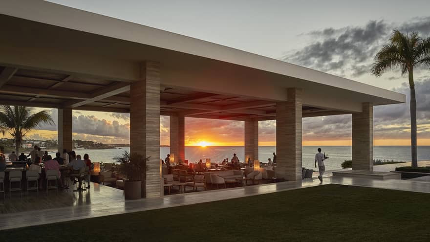 Open-air seaside restaurant with guests dining at sunset, overlooking the ocean.
