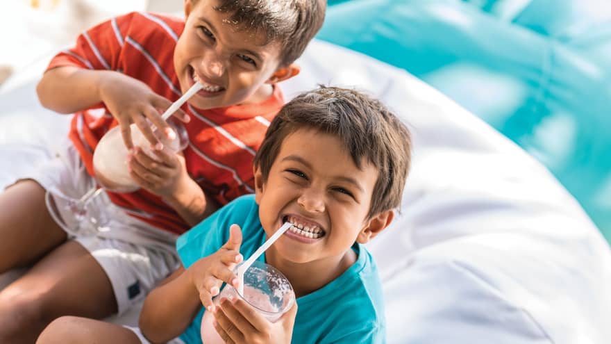 Two boys smile really big smiles as they sip on a virgin piña colada on a bean bag cushion