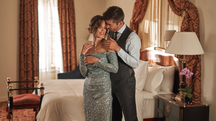 Couple in black tie attire embrace in front of bed in guest room