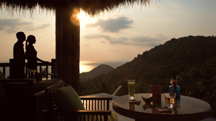 Silhouettes of man and woman, cocktail table on patio overlooking mountains at sunset