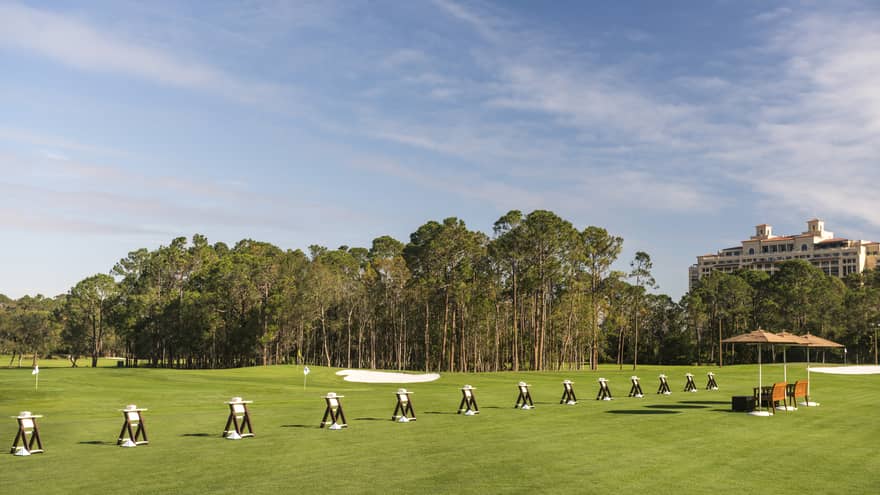 Driving range set up alongside a putting green with three orange umbrellas nearby and a tree line in the distance