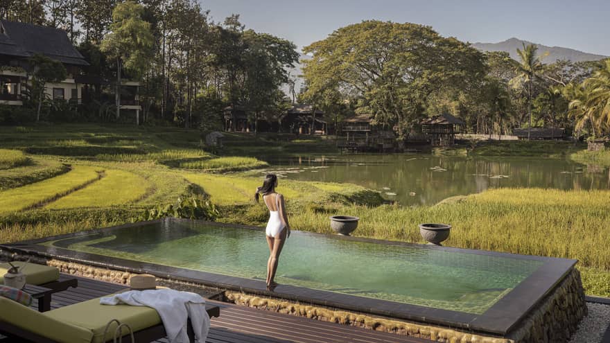Person standing at the edge of an infinity pool overlooking rice fields, a tranquil pond and forested hills.