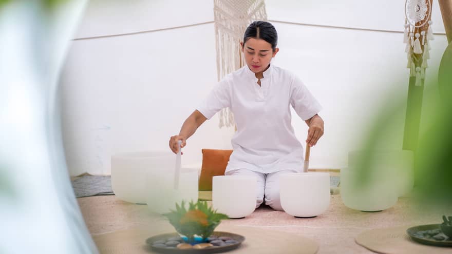 A person in white performs a healing ritual with crystal bowls inside a tranquil wellness tent, surrounded by soft décor and greenery
