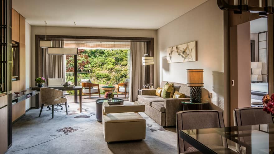 Living area with sofa, cushions, coffee table, armchair, marble desk and a private balcony with outdoor seating overlooking a lush garden