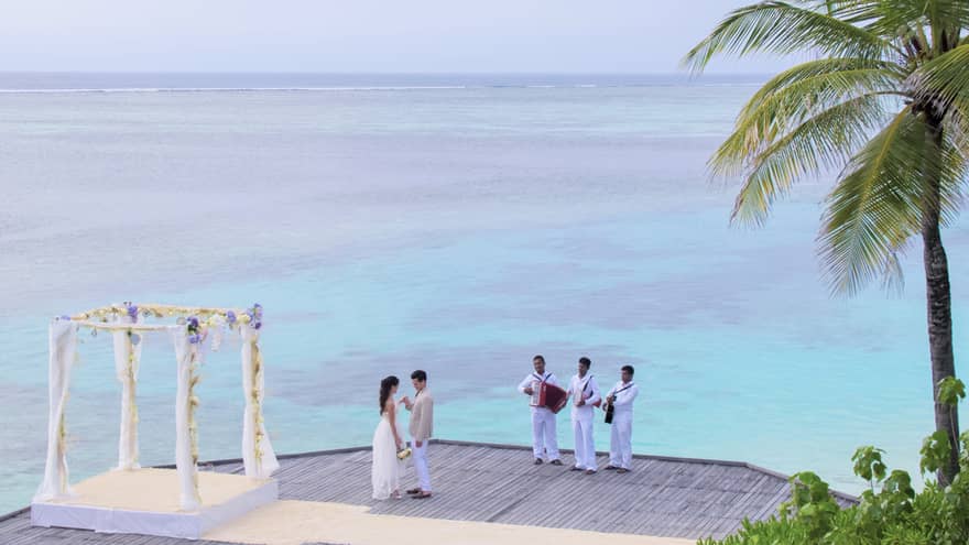 Wedding ceremony with bride and groom, three musicians on dock over lagoon, palm tree