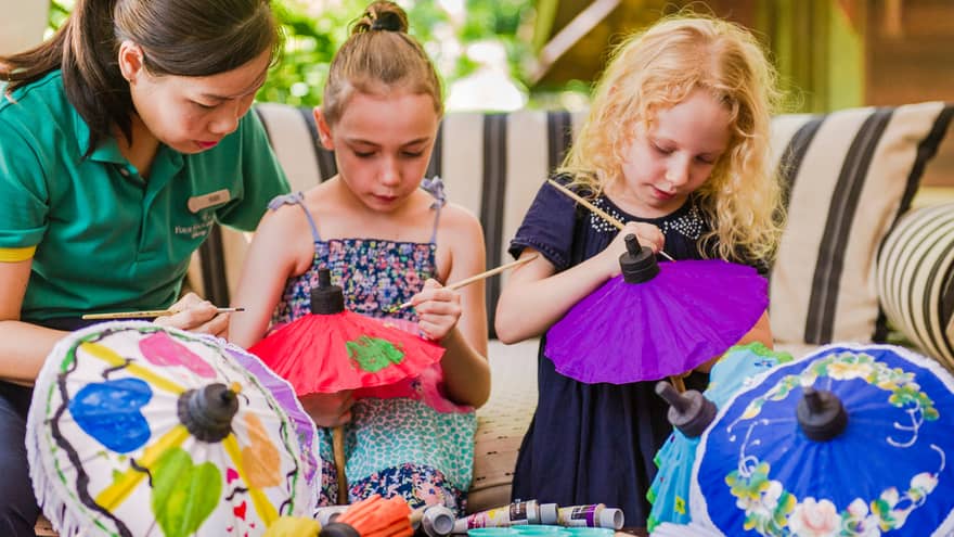 Kids Club staff helps young girls paint pictures on colourful paper umbrellas
