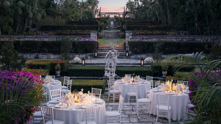 Round white banquet tables with candles on stone patio under hedges