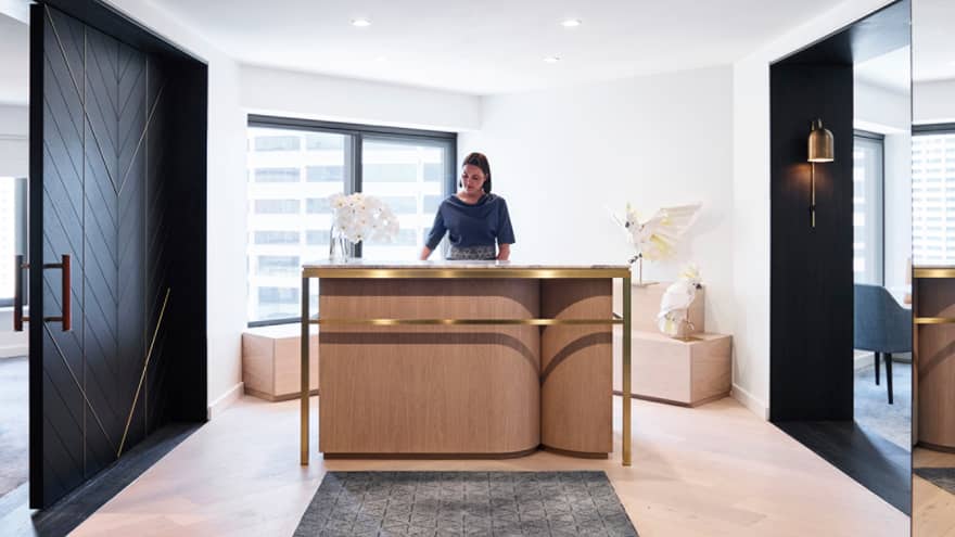Hotel hostess stands at modern reception desk by window, meeting room doors at end of long hall