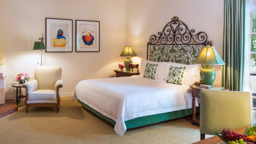 Jungle Room bed with iron and green-padded headboard, white armchair by fireplace, desk, fruit bowl